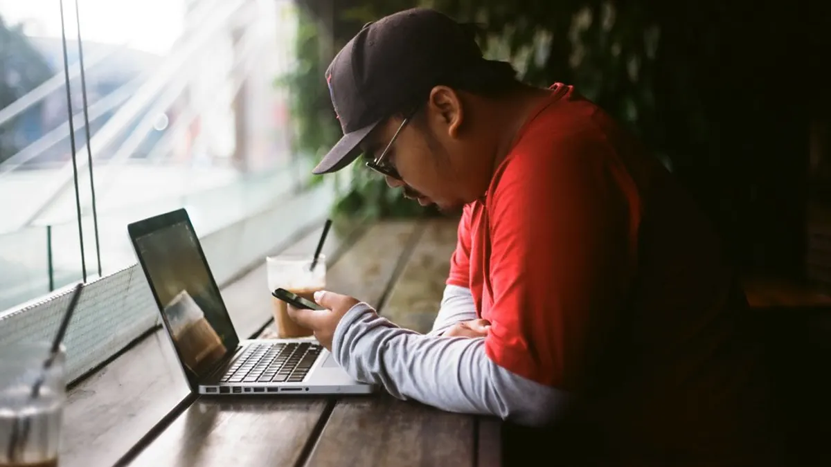 Man kijkt op zijn telefoon aan een houten tafel met een laptop en een koffie