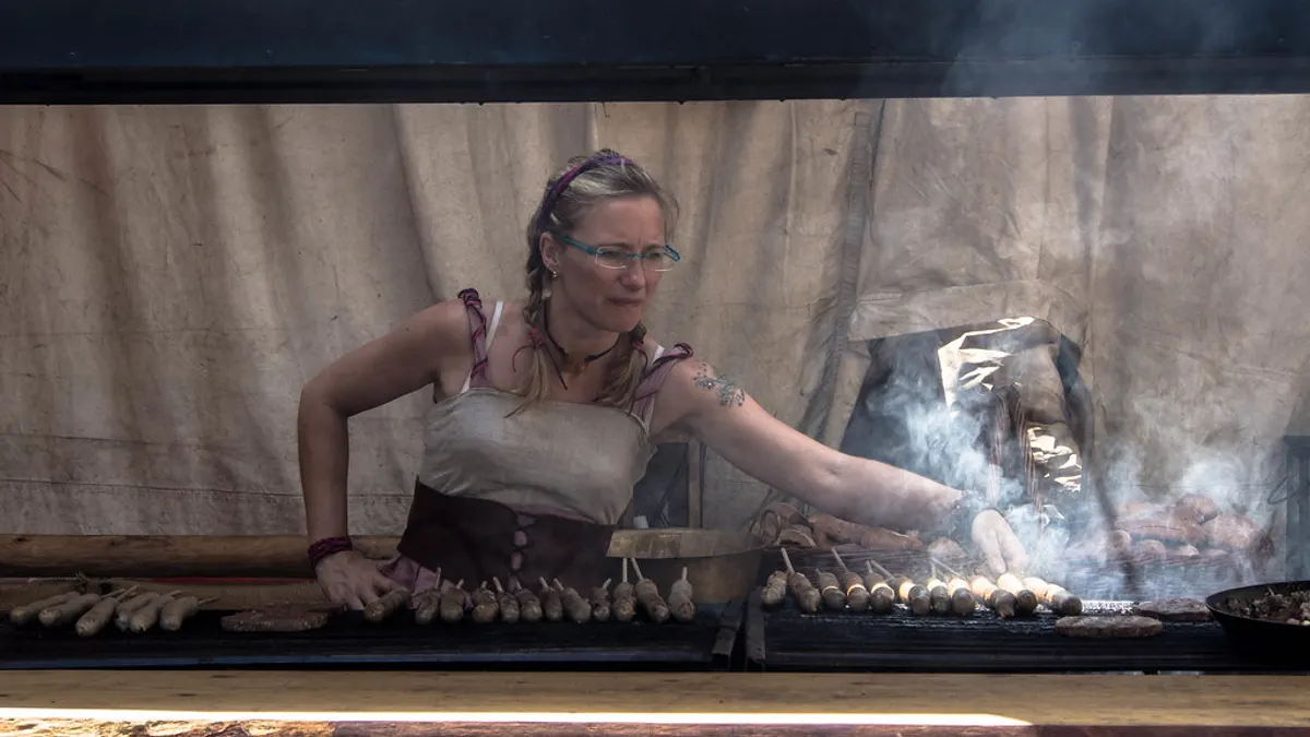 Een kok bakt rijen worstjes en spiesen op een lange houtskoolgrill met rook boven het vuur