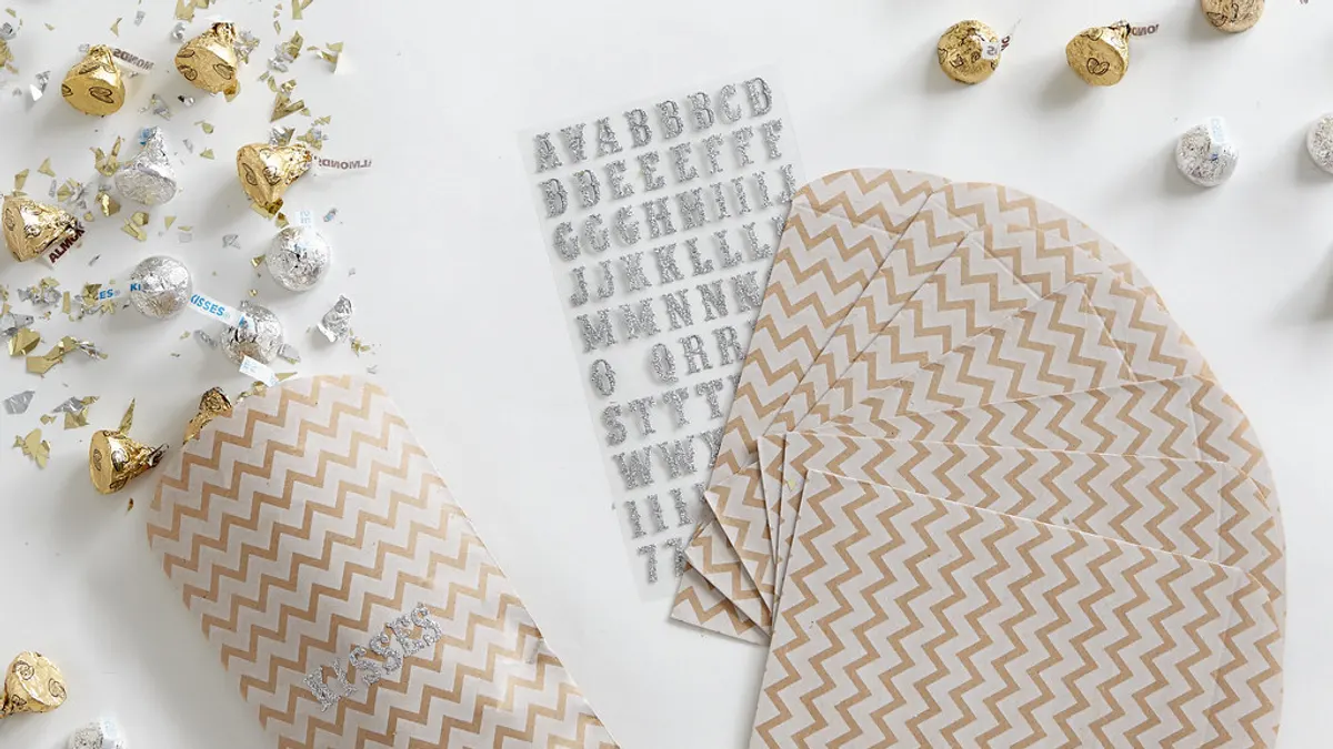 Papieren zakjes met een goudkleurig zigzagmotief, een vel letterstickers en losse chocolaatjes in folie op een witte tafel
