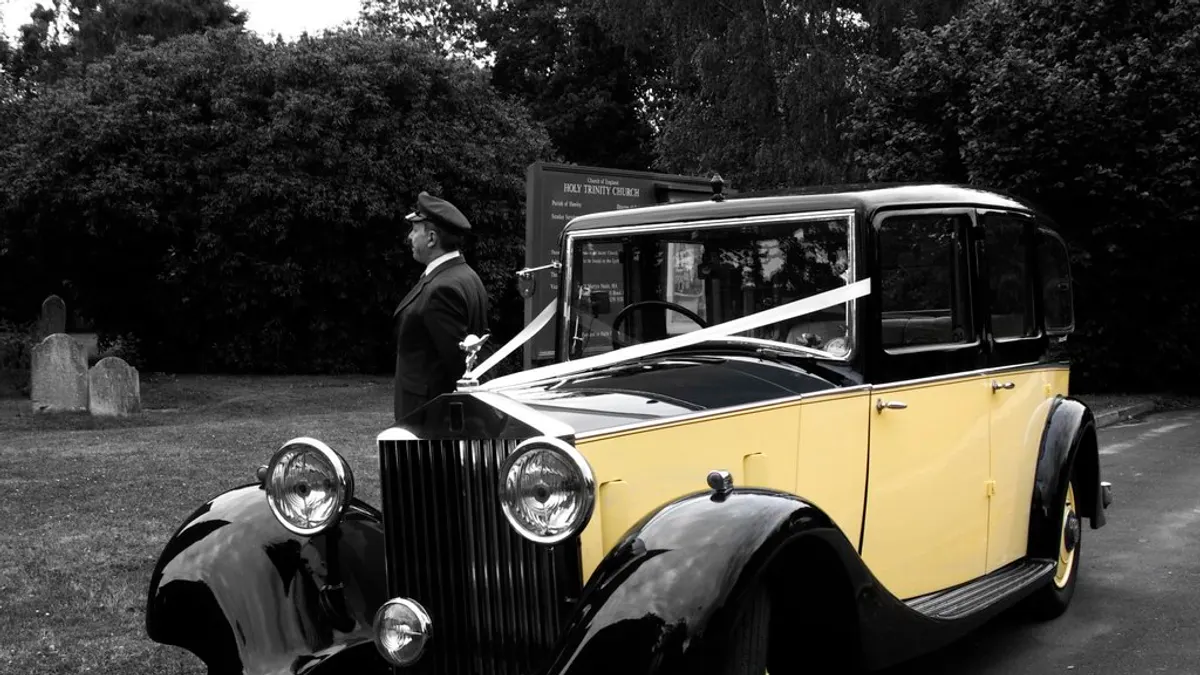 Klassieke oldtimer met wit trouwlint en een chauffeur ernaast in zwart-wit