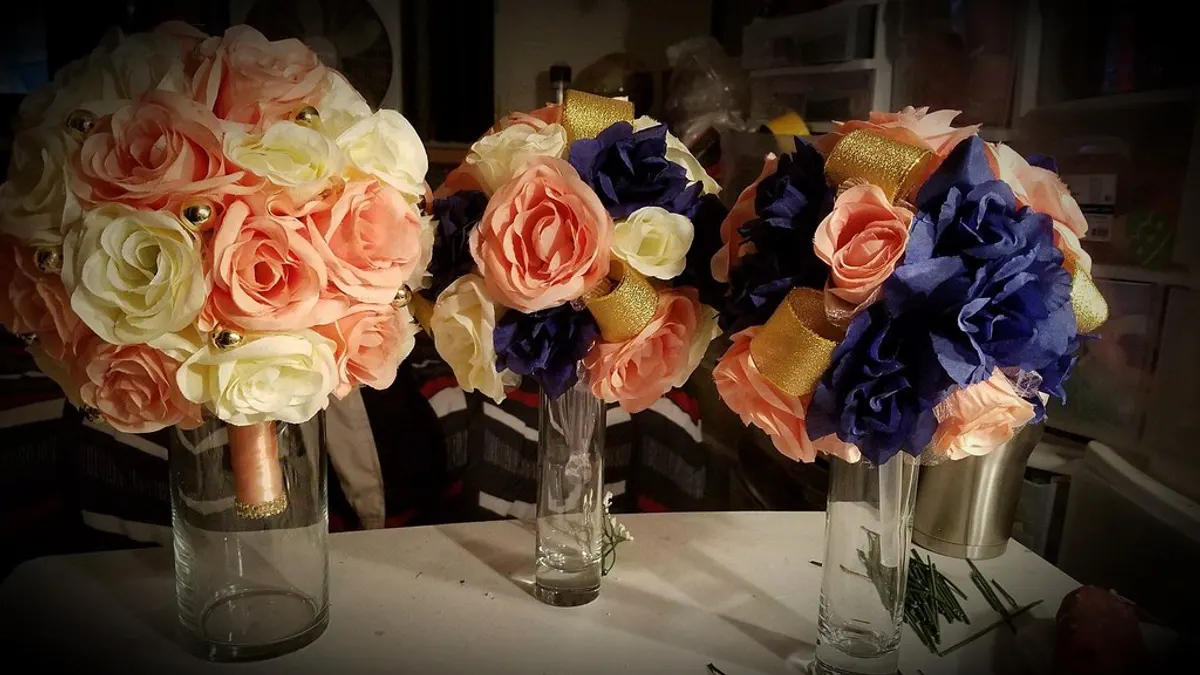 Een groot bruidsboeket naast twee kleinere bruidsmeisjesboeketten met perzikroze en blauwe bloemen in glazen vazen