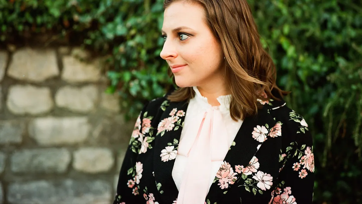 Vrouw in een zwarte blazer met roze bloemenprint en een lichte blouse met strik, voor een begroeide muur