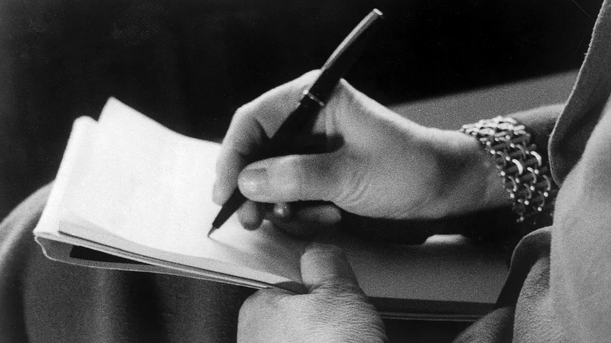 Zwart-witfoto van een hand met een armband die met een pen een bericht schrijft in een notitieboekje