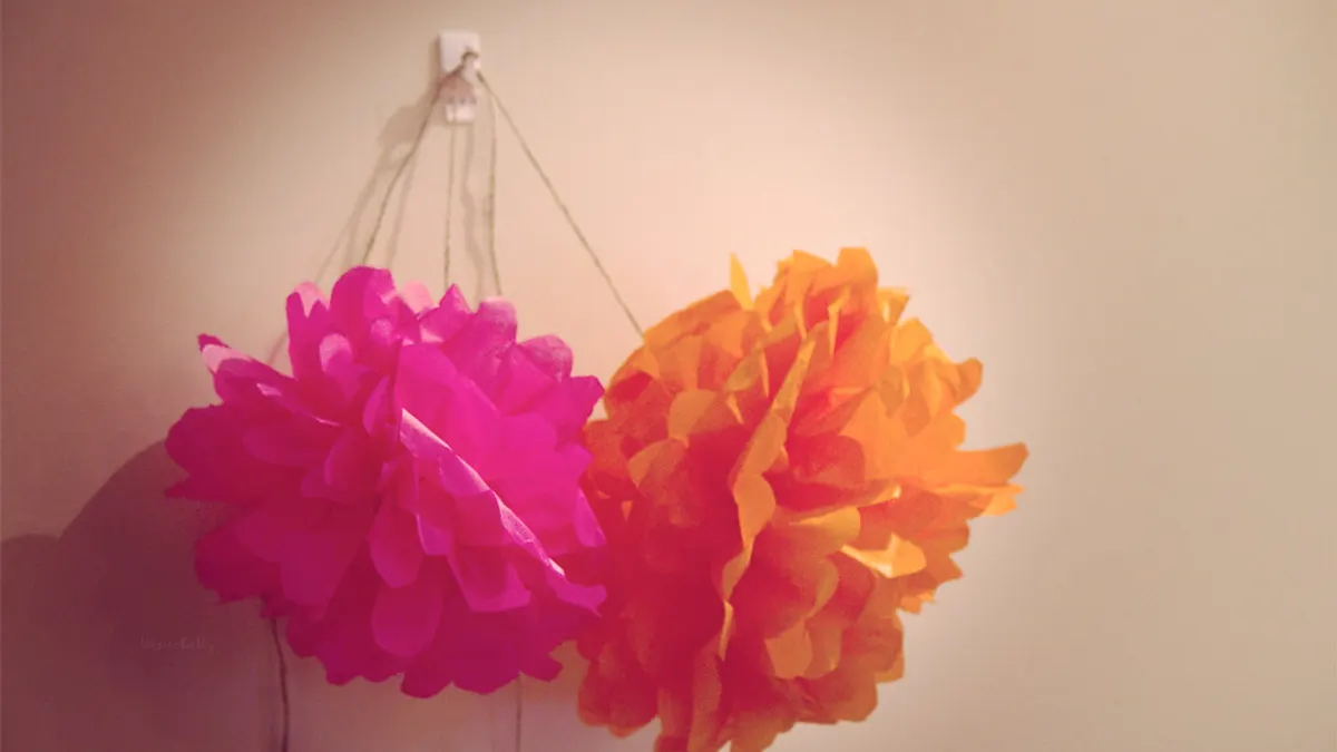 Twee papieren pompons in fuchsia en oranje hangen aan een muur