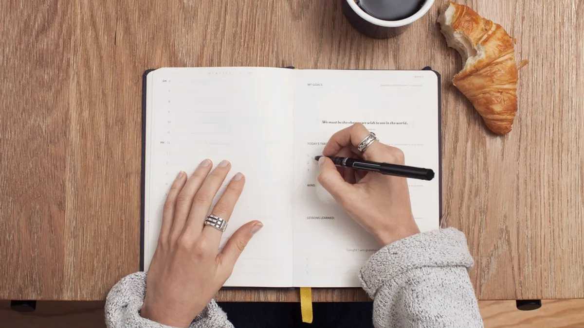Iemand schrijft in een opengeslagen planner op een houten tafel, met een kop koffie en een croissant ernaast