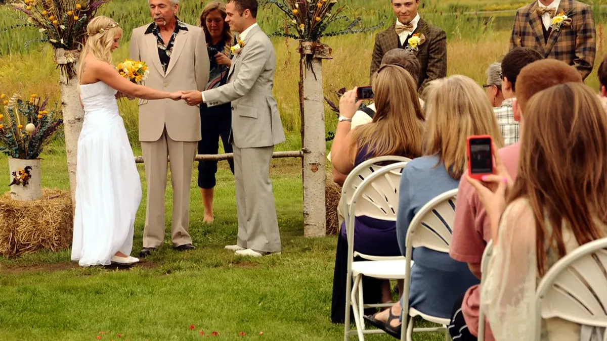 Een trouwambtenaar staat bij het bruidspaar tijdens een ceremonie in open lucht, met gasten op witte stoelen