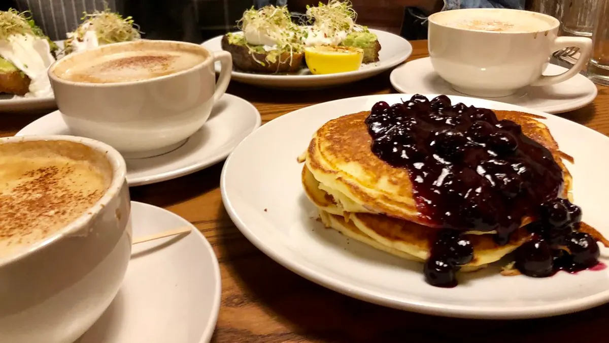 Een brunchtafel met grote koppen cappuccino, toast met avocado en een stapel pannenkoeken met blauwe bessen