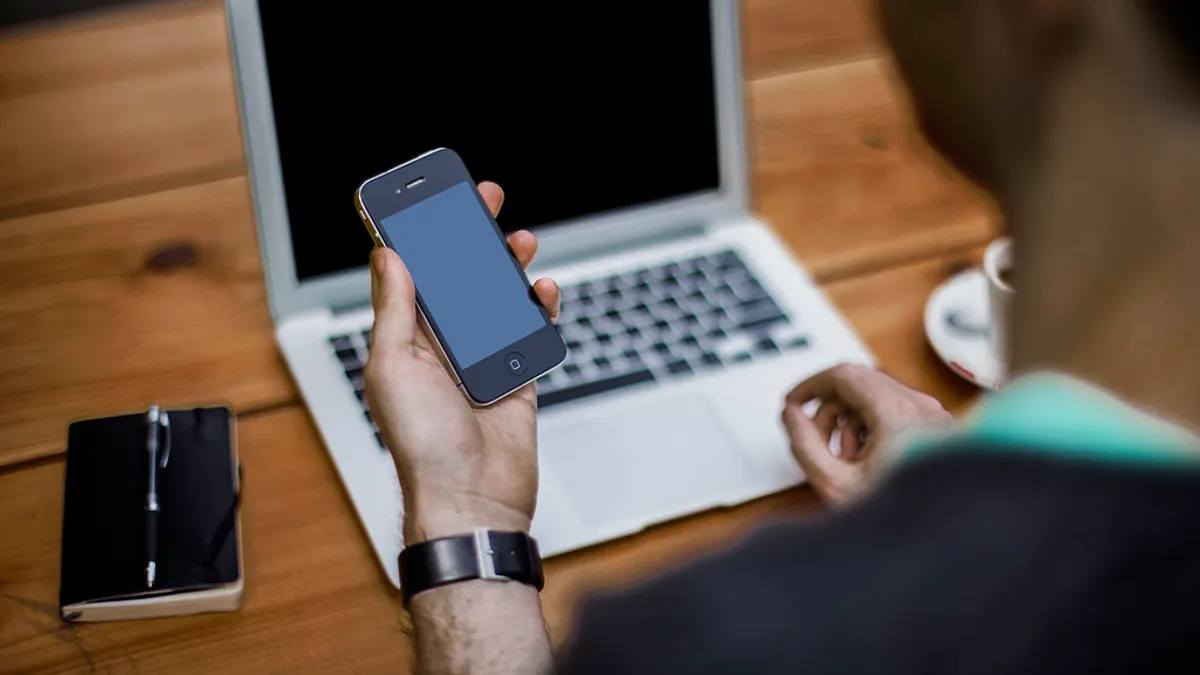 Hand met een smartphone aan een houten tafel, met een laptop en een notitieboekje ernaast