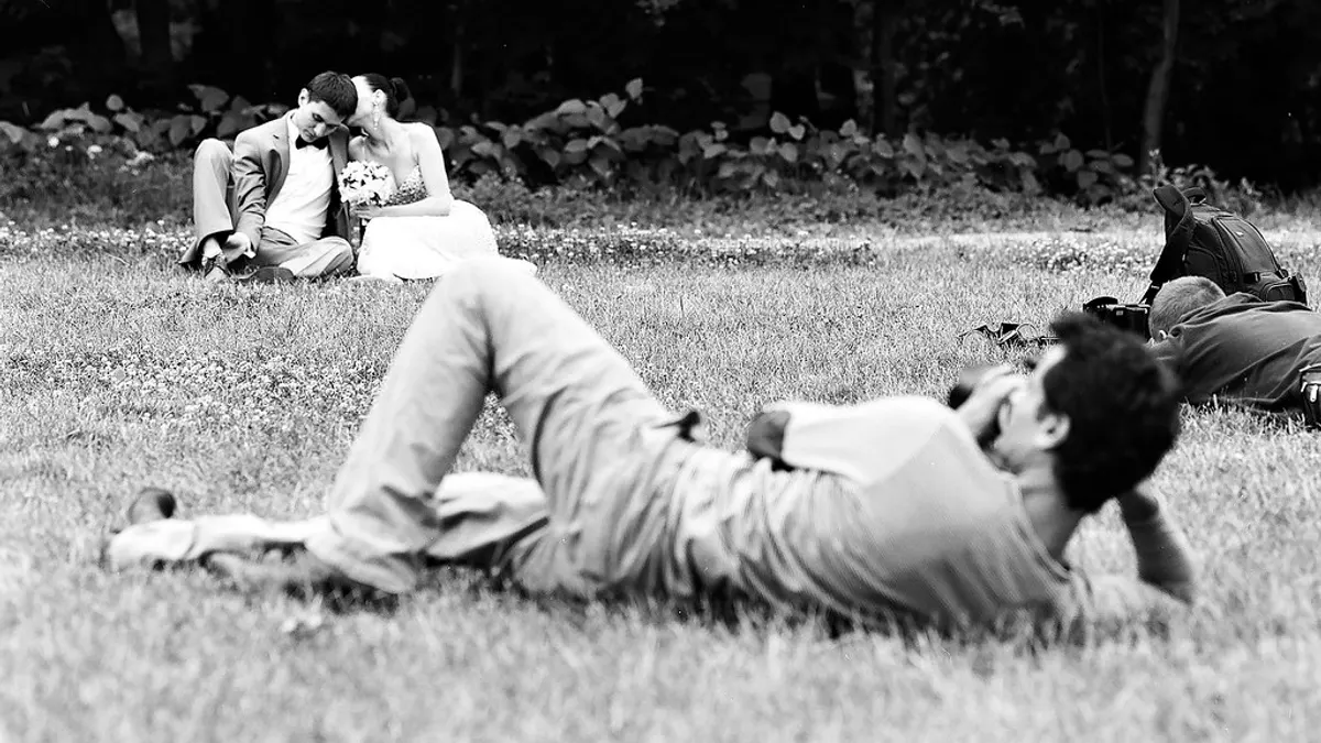 Een fotograaf ligt in het gras en fotografeert het bruidspaar, een tweede fotograaf ligt verderop met zijn camera klaar