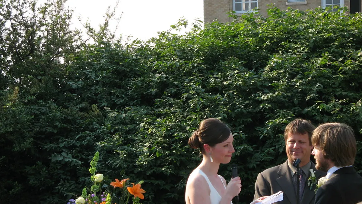 Bruid spreekt haar trouwbelofte uit in een microfoon tijdens een buitenceremonie, met de officiant en de bruidegom naast haar