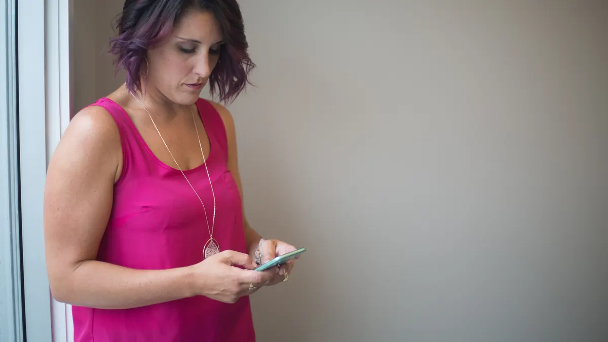 Een vrouw in een roze topje typt bij een raam een bericht op haar telefoon