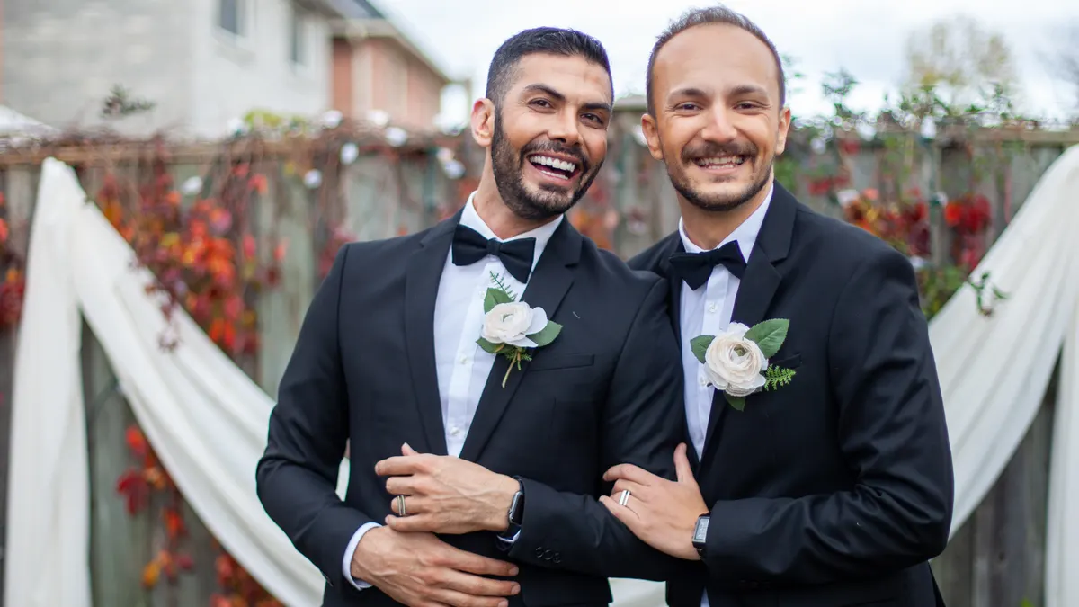 Twee lachende bruidegoms in smoking met een witte corsage, arm in arm voor een versierde schutting