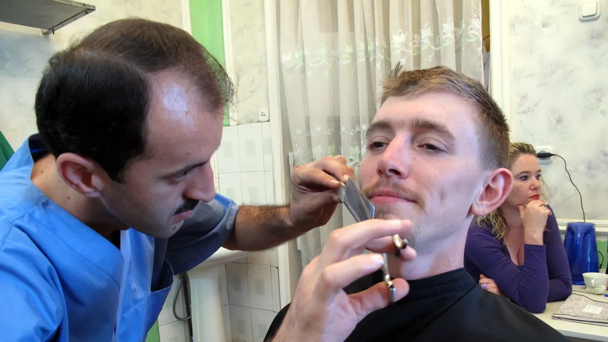 Kapper werkt met kam en schaar de baard bij van een man in een kappersmantel