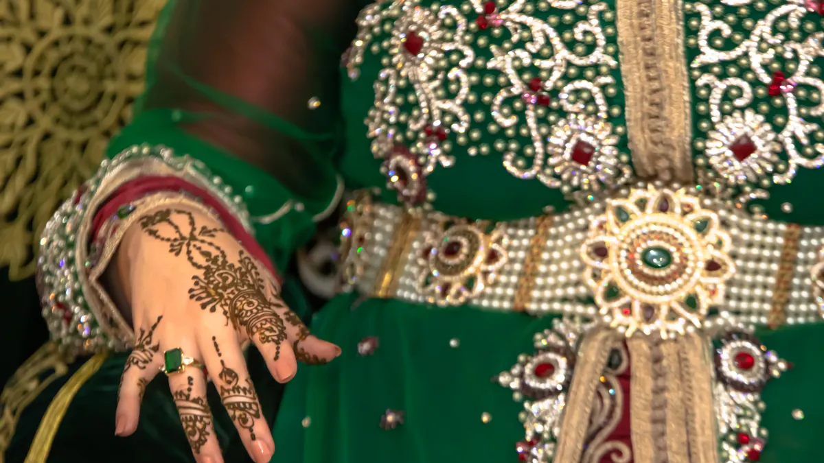 Hand met hennaversiering naast een groene kaftan met parels, stenen en een rijk versierde riem