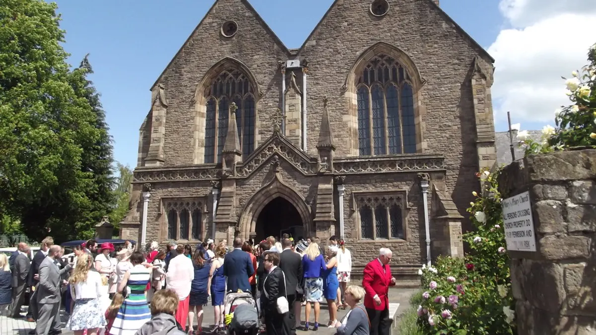 Drukte van trouwgasten in zomerse kleding voor een stenen kerk op een zonnige dag