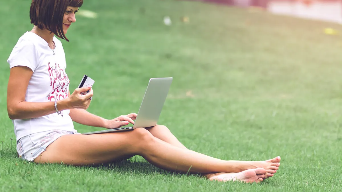Vrouw zit in het gras met een laptop en houdt een bankkaart vast om online te betalen
