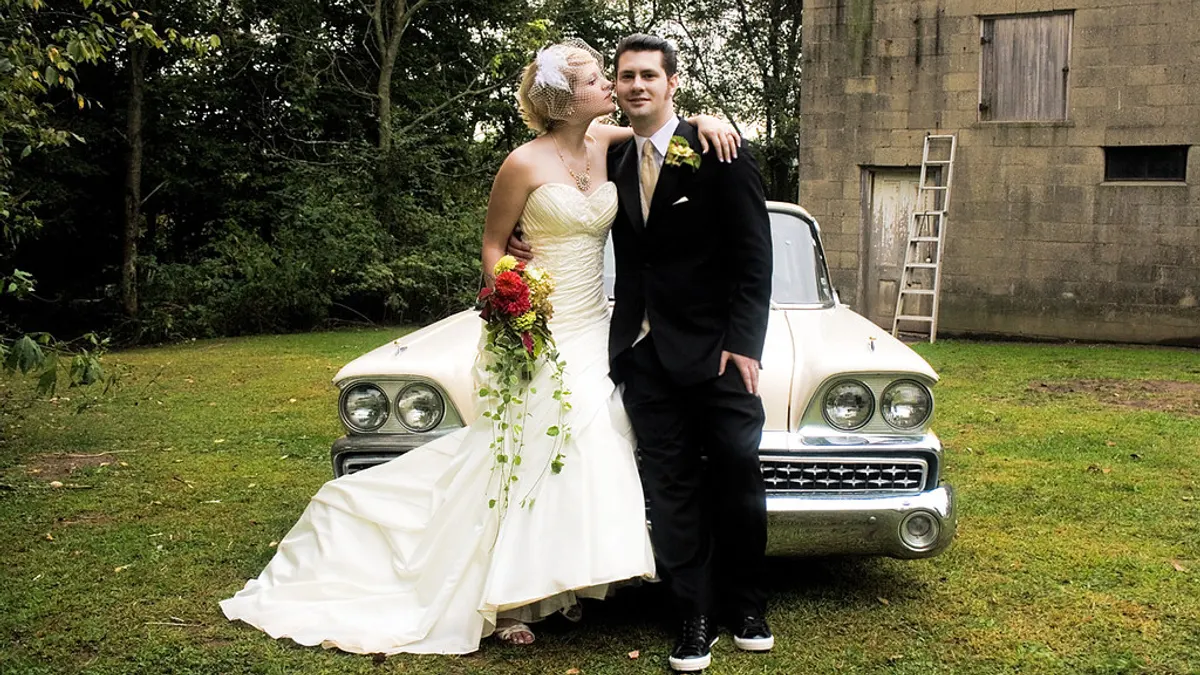 Een bruidspaar poseert op het gras voor een witte oldtimer uit de jaren vijftig