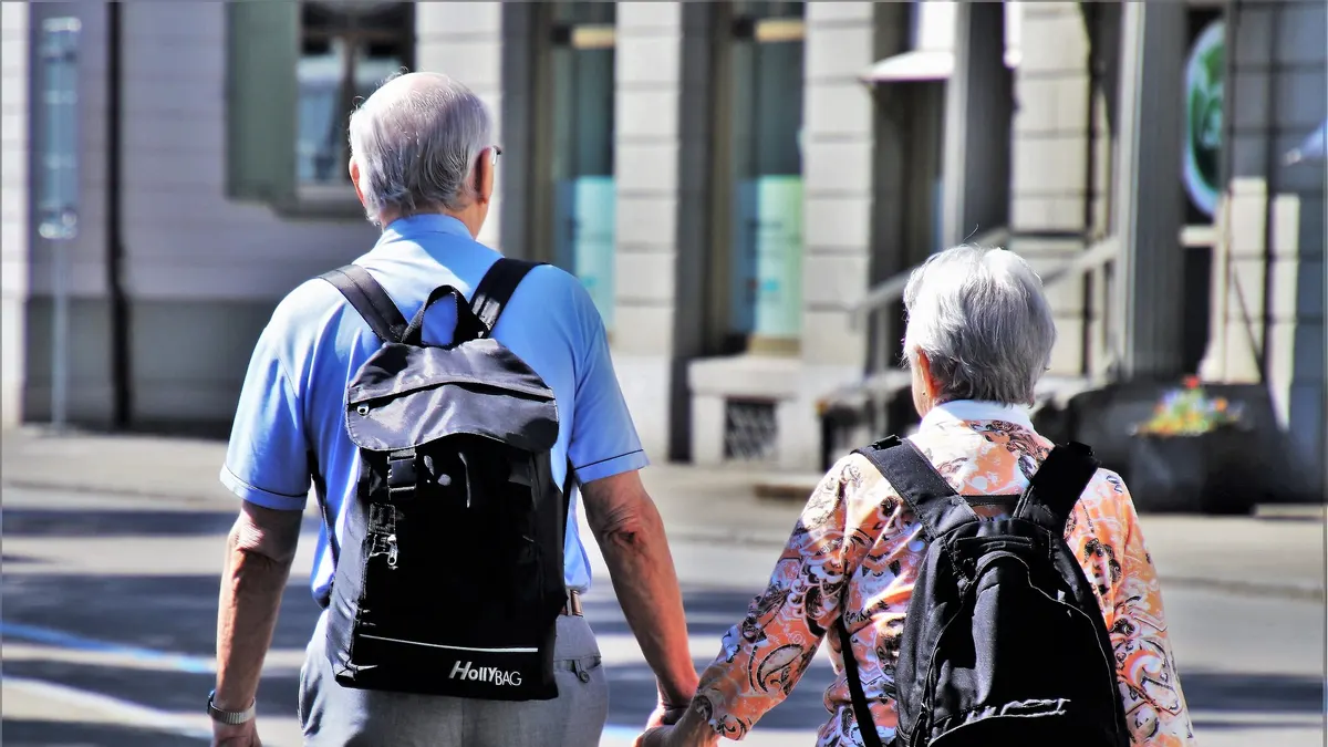 Een ouder koppel met rugzakken wandelt hand in hand door een zonnige straat, van achteren gezien