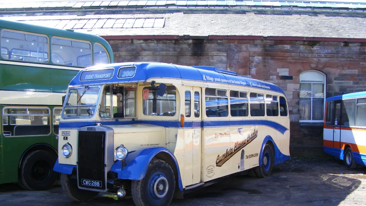Een crème met blauwe oldtimerbus staat geparkeerd voor een stenen remise