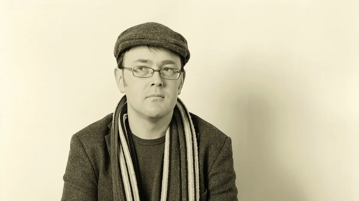 Man met een tweed flatcap, visgraatjasje en gestreepte sjaal in sepia voor een lichte muur