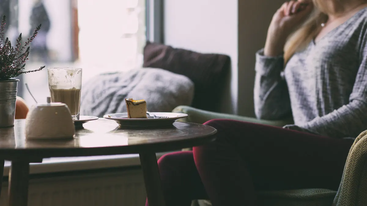 Vrouw zit in gedachten bij het raam, met een koffie en een stuk taart op het tafeltje naast haar