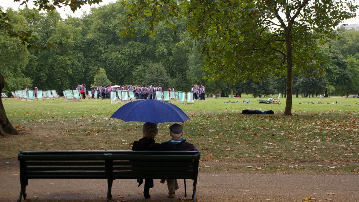 Een ouder koppel zit samen onder een blauwe paraplu op een bank in een regenachtig park
