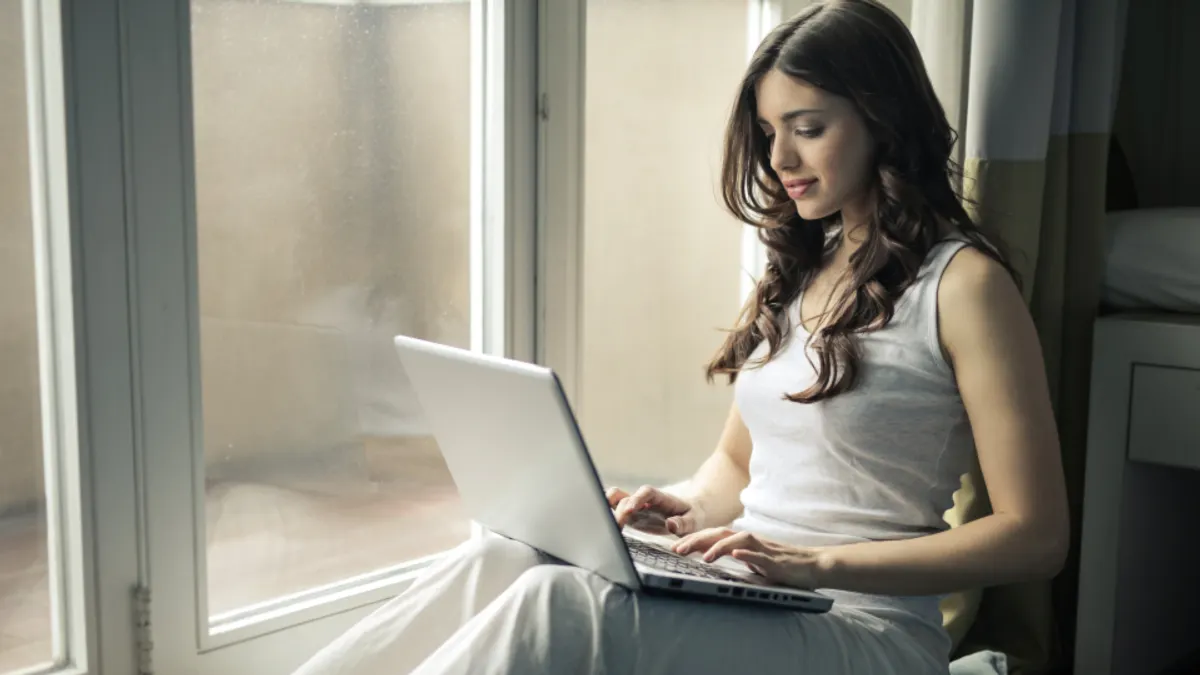 Een vrouw zit bij het raam en werkt op een laptop op haar schoot
