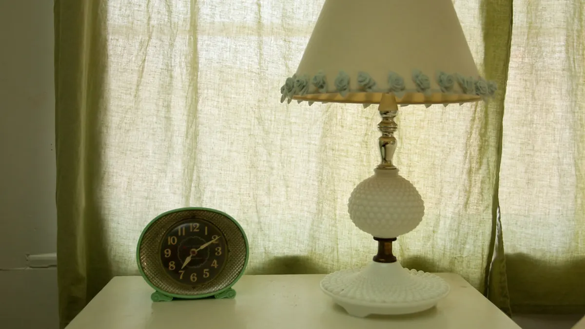 Een nachtkastje met een ouderwetse groene wekker en een witte lamp voor een licht gordijn
