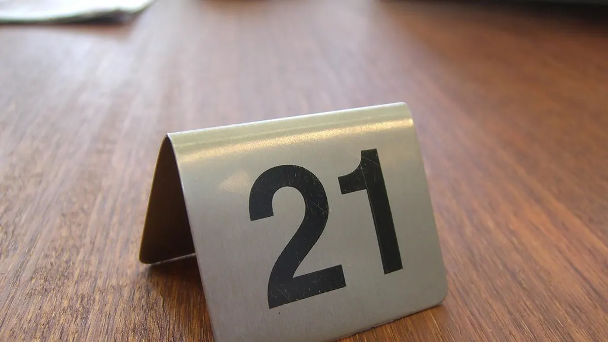 Een metalen tafelnummer 21 in grote zwarte cijfers op een houten tafelblad