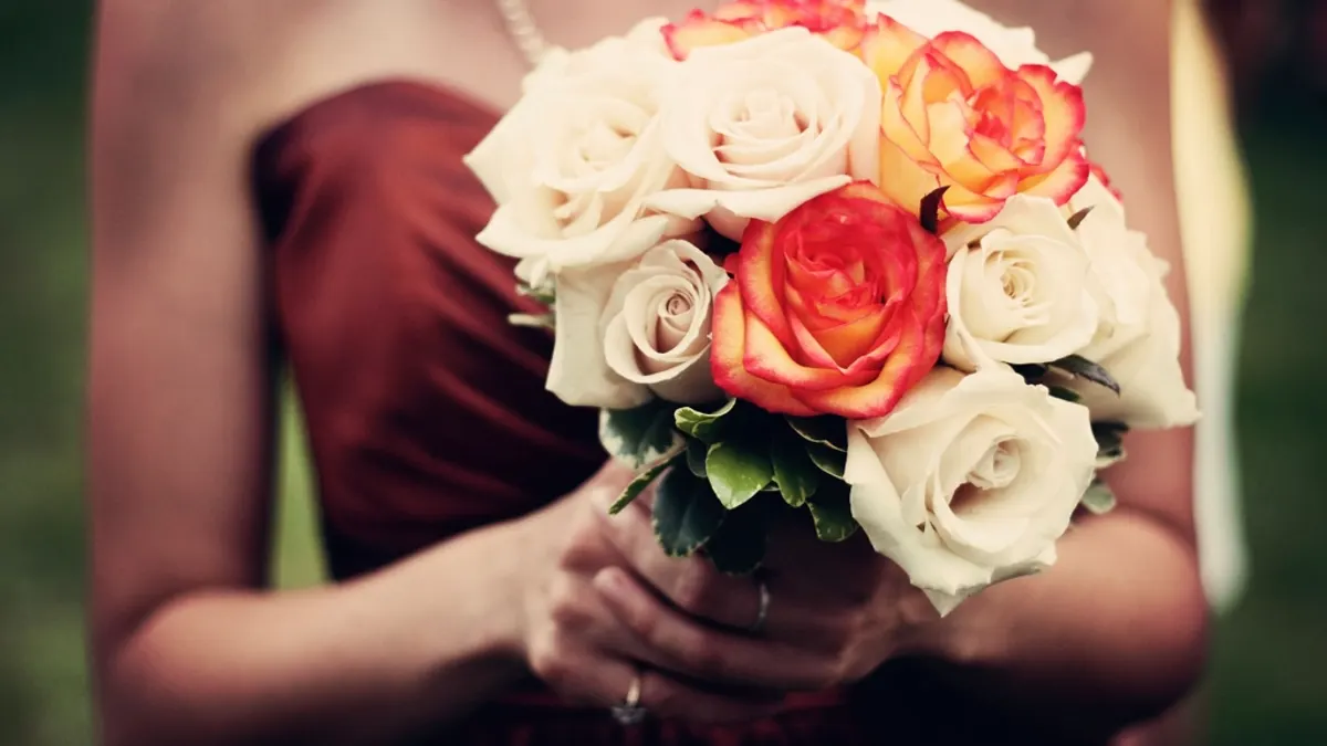 Bruidsmeisje in een donkerrode jurk houdt een boeket witte en oranje rozen met beide handen vast