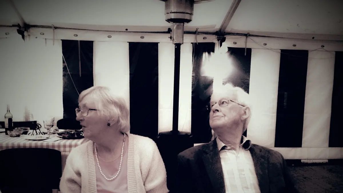 Een ouder koppel zit aan tafel onder een feesttent met een terrasverwarmer achter hen