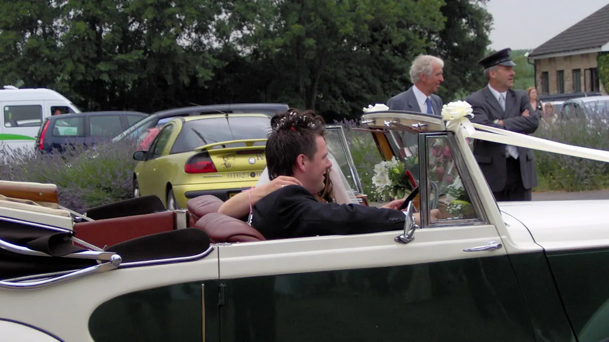 Bruidegom stapt in een witte cabriolet-oldtimer terwijl de chauffeur toekijkt