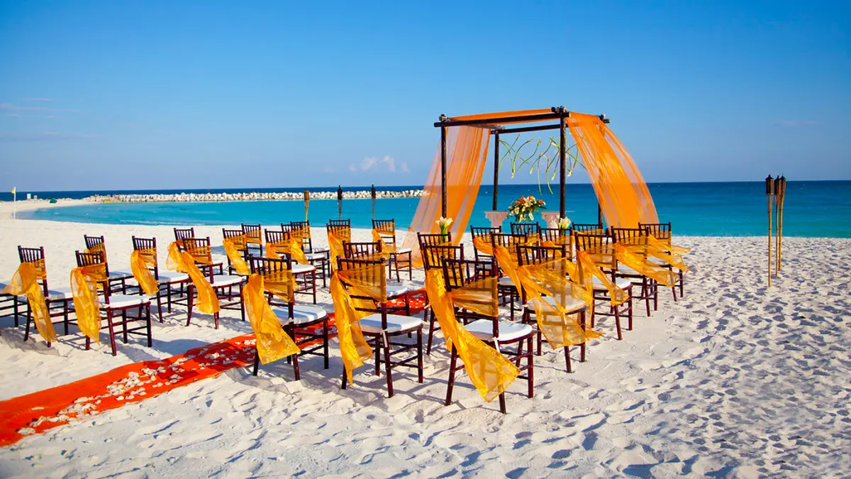 Stoelen met oranje linten en een prieel klaar voor een ceremonie op een wit strand aan een turquoise zee
