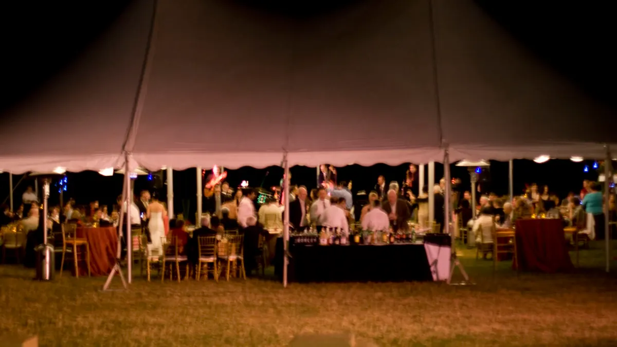 Verlichte feesttent op een gazon met gedekte tafels en gasten op een avondfeest
