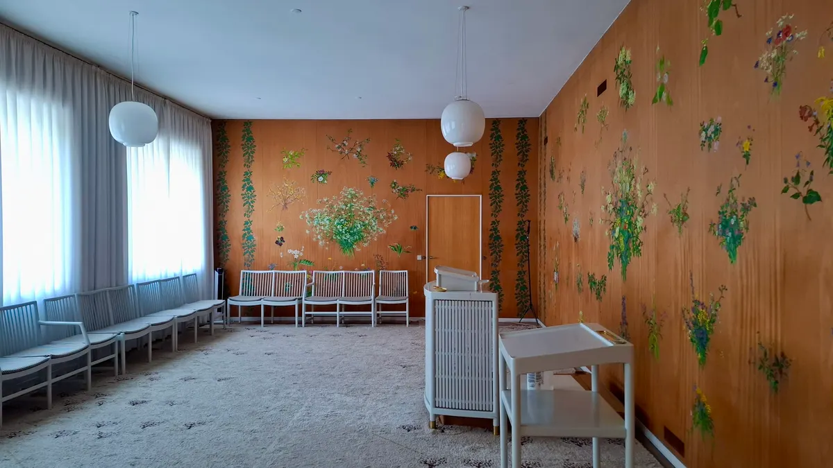 Een lichte trouwzaal in een stadhuis met witte stoelen langs de wanden, een spreekgestoelte en beschilderde houten panelen met bloemen