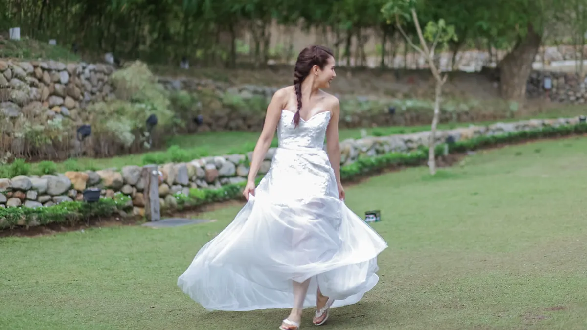 Bruid met een vlecht in een strapless jurk met glanzend lijfje en wijde tulerok wandelt over het gras van een tuin