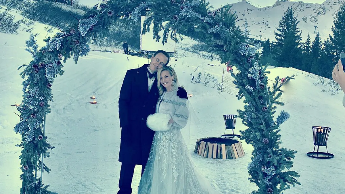 Bruid in een kanten jurk met lange mouwen en een witte bontmof staat met de bruidegom onder een boog van dennengroen in de sneeuw