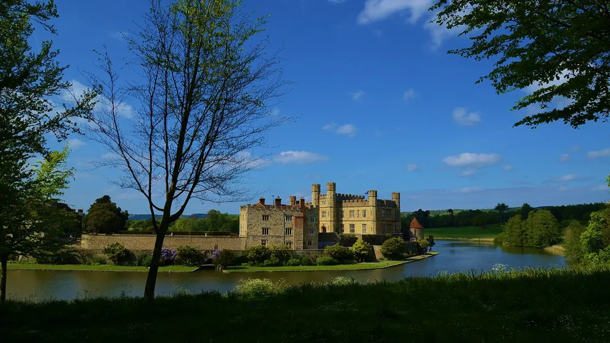 Kasteel aan een brede slotgracht, omringd door gazon en hoge bomen onder een blauwe lucht