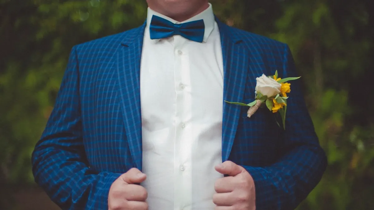Bruidegom in een blauw geruit colbert met strik en een corsage van een witte roos, de handen aan de revers