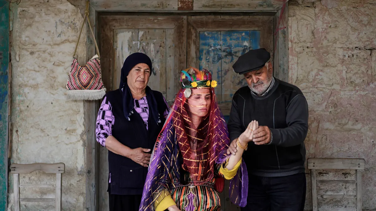 Een bruid in traditionele Turkse kleding met een rode sluier krijgt een gouden armband om van een oudere man, naast een oudere vrouw