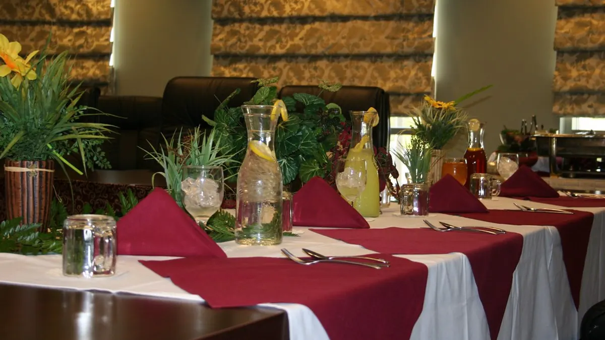 Gedekte tafel met bordeaux servetten, karaffen limonade en groene planten, klaargezet voor een feest