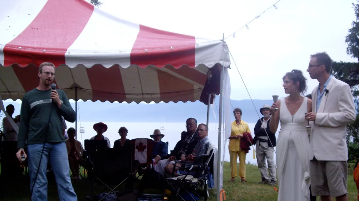 Een spreker met microfoon staat buiten bij een feesttent op het gras, naast het bruidspaar en de zittende gasten