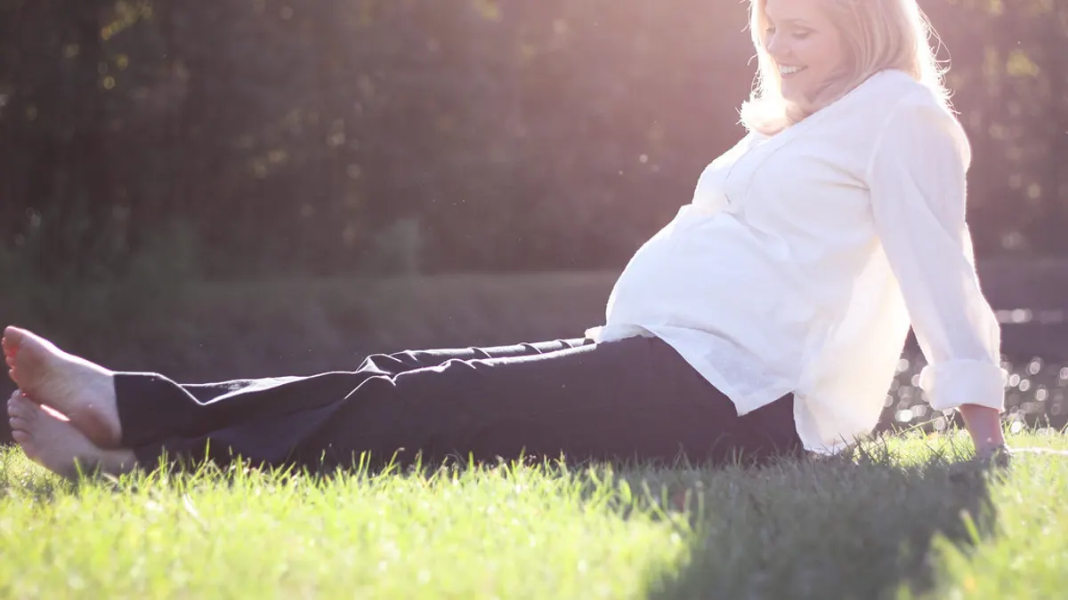 Een zwangere vrouw in een losse witte blouse en een zachte broek zit ontspannen in het gras in de avondzon